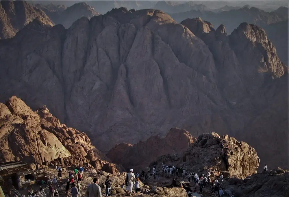 people climbing a desert mountain of arid rock with a towering steep rock mountain in the background