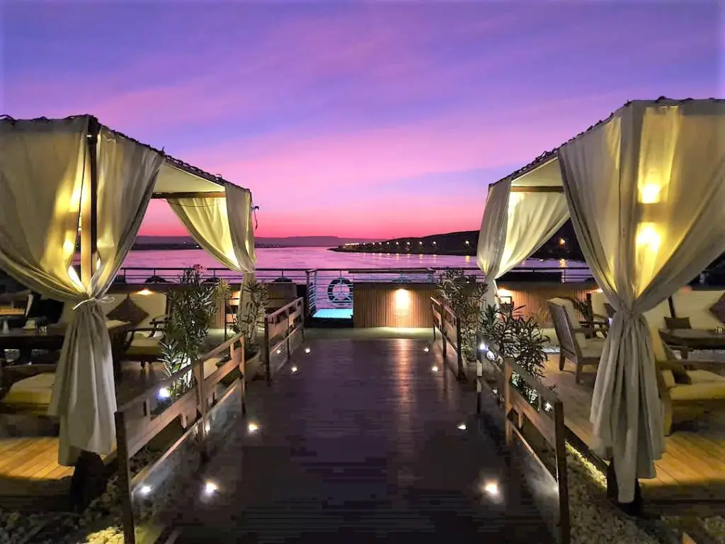 A cruise ship on the Nile shows seating on the upper deck with hanging curtains and seating and a pink sunset in the background