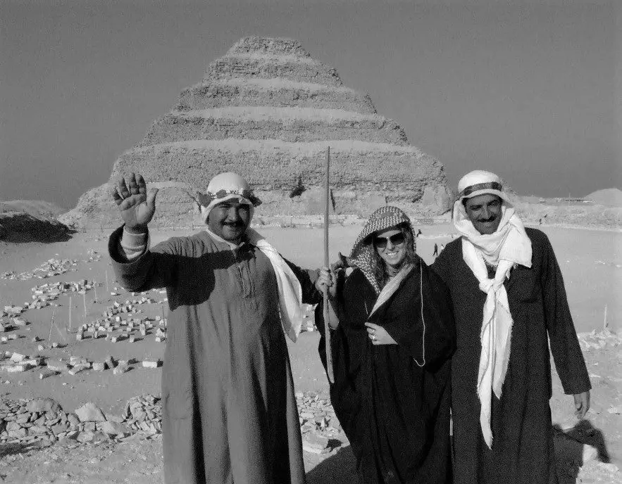 Standing next to 2 Egyptian men. We are all dressed in robes with the Step Pyramids behind us.