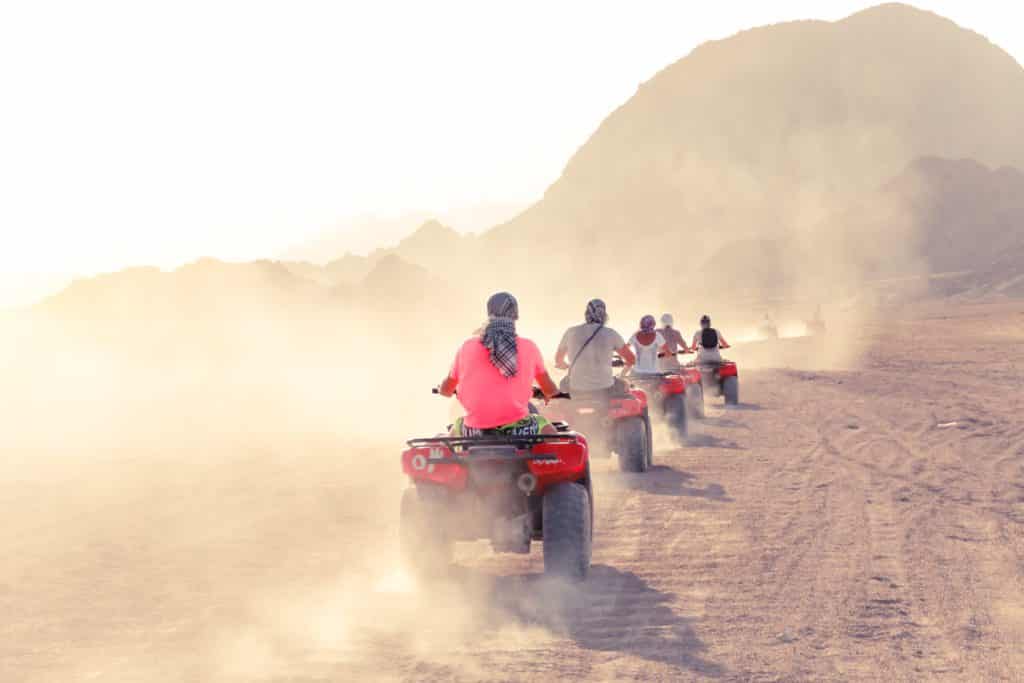 A row of 5 quad bikes going through the desert. Dust rising from the bikes.