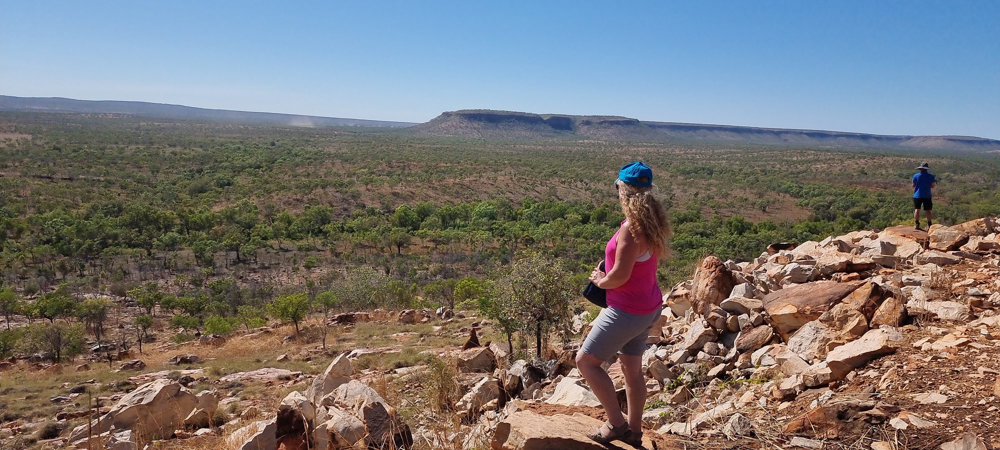 a lady wearing a pink tp and grey shorts  observes the King leopold Ranges. The green trees and mountains in the background are set against a blue sky